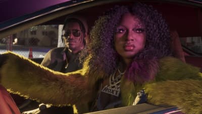 A woman in a yellow fur coat and a diamond Roxy nameplate rides in a car's passenger seat at dusk.